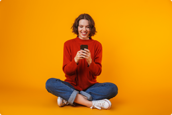 Kobieta na żółtym tle siedząca po turecku i trzymająca w dłoni telefon z aplikacją.